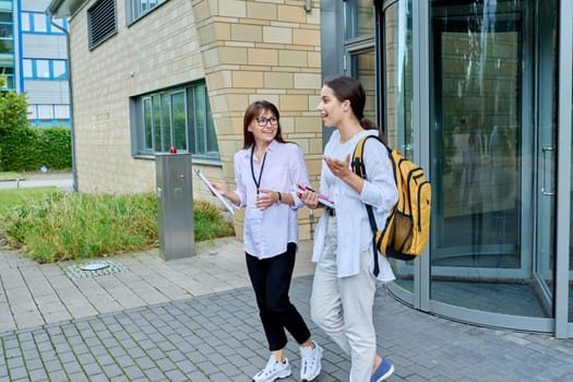 Teenage girl high school student with backpack talking to female teacher, mentor, coach, standing outdoors on educational building background. Adolescence, education, knowledge, communication