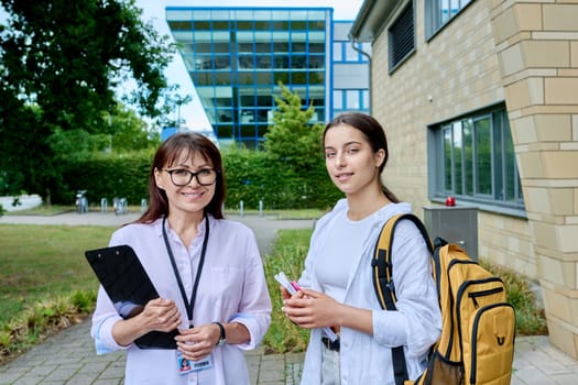 Teenage girl high school student with backpack talking to female teacher, mentor, coach, standing outdoors on educational building background. Adolescence, education, knowledge, communication