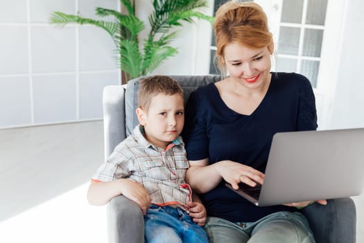 Woman with boy working on laptop online on armchair