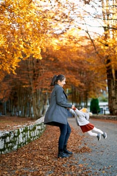 Mom spins a little girl by the arms while standing on the road in the autumn park. High quality photo