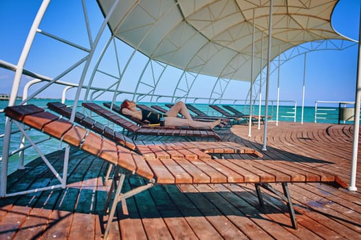 Woman sunbathing in lounge chair on wooden pier on sea. Beach vacation, summer leisure and travel