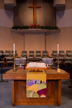 Tranquil Worship: A serene church interior in Fort Wayne, Indiana, showcases a wooden pulpit adorned with a colorful landscape, an open Bible, and elegant candles. A large wooden cross, accented by a decorative wreath on back wall.
