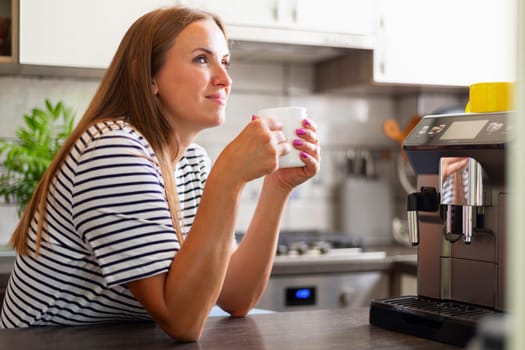 Young woman enjoying freshly prepared aromatic coffee near modern coffee machine in kitchen.