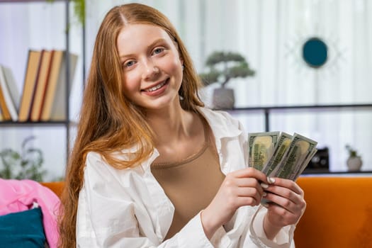 Happy rich business teenager red hair child girl cheering up with high money profits, lottery game win, satisfied of wealth income earnings salary sits on home room couch full of dollar cash banknotes