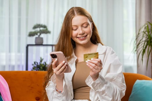 Teenager girl using credit bank card and smartphone while transferring money, purchases online shopping cashless, order food delivery at home apartment indoors. Happy child sitting in room on couch