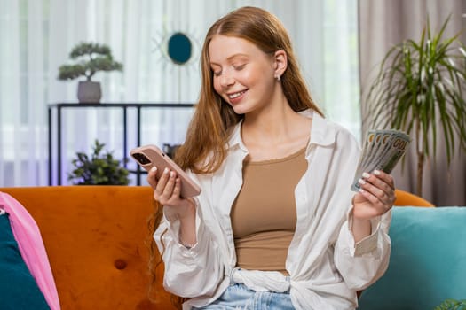 Planning family budget. Smiling teenager girl counting money cash, use smartphone calculate domestic bills at home room. Joyful child satisfied of income and saves money for planned vacation, gifts