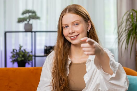 Hey you. Young teenager girl smiling excitedly and pointing to camera, choosing lucky winner, indicating to awesome you, inviting approve. Portrait of child at modern home living room sitting on couch