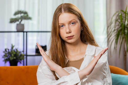 Stop gesture. Angry teenager red hair girl say No hold palm folded crossed hands, warning of finish, prohibited access, declining communication, body language, danger. Child at home room sits on sofa