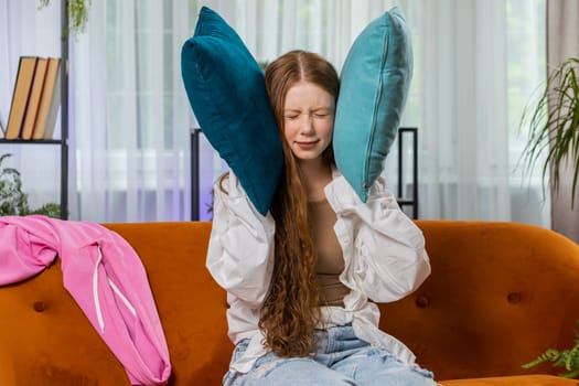 Repair work at neighbours. Irritated girl child relaxing on couch cover ears with pillows annoyed by noisy neighbors suffer from headache wish silence. Thin walls at home flat without sound insulation