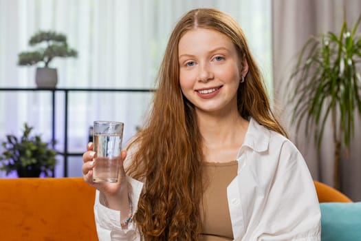 Thirsty teenager girl holding glass of natural aqua make sips drinking still water preventing dehydration at home sitting on sofa. Child with good life habits, healthy slimming, weight loss concept