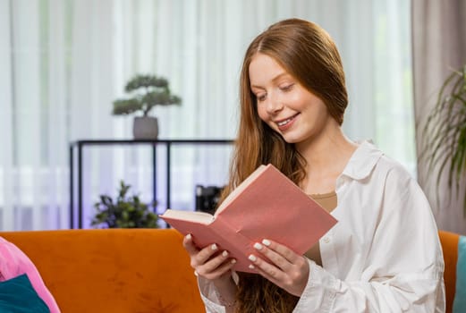 Teenager red hair girl relaxing reading interesting book, turning pages smiling enjoying literature, taking a rest on comfortable couch. Portrait of happy child at home apartment living room on sofa