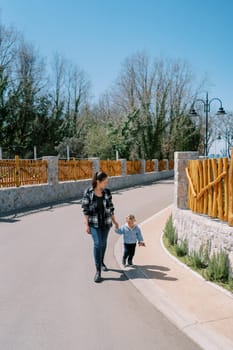 Mom and a little girl walk holding hands along the asphalt road along the rural fences. High quality photo
