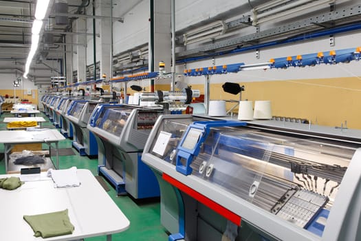 A row of industrial textil flat knitting machines in a knitwear factory. An industrial line of modern automatic knitting machines arranged in two rows.