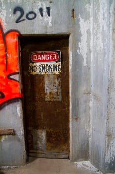 Decaying industrial doorway in East of St. Louis, Illinois. Rusty metal door adorned with weathered 'DANGER NO SMOKING' sign hints at hazardous conditions. Urban decay and cautionary themes.