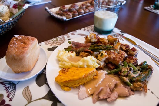 Indulge in a festive feast with a traditional American spread. A heaping plate of mashed potatoes, roast turkey, cornbread, bacon-wrapped asparagus, and green bean casserole, accompanied by a roll.