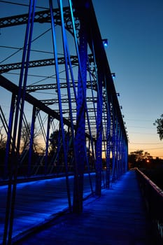 Twilight serenity on the Wells Street Bridge, Fort Wayne, Indiana, as blue lights illuminate the stunning steel truss structure against a backdrop of warm sunset hues.