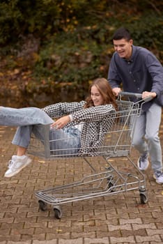 Autumn Joyride: Boy Rolling Girl in Shopping Cart amid Nature's Beauty. playful couple, autumnal adventure, outdoor fun, seasonal joy, laughter and love, romantic play, nature's embrace, joyful moments, couple goals, autumn magic.