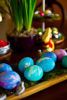 Vibrant Easter eggs decorated in shades of blue, purple, and pink, nestled on moss and displayed on a rustic wooden tray. A festive scene with additional decorations and green plants in the background.