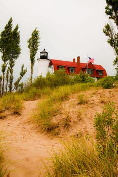 Iconic lighthouse stands tall against overcast sky, its black lantern room guiding ships to safety. Vibrant red-roofed building and waving American flag evoke a sense of coastal patriotism. Serene beach grass-lined path adds depth.