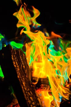 Vibrant and mesmerizing, this close-up shot showcases a campfire's fierce orange flames and intriguing green hues, creating a mystical ambiance. The charred wood and glowing embers add to the dynamic energy, making it perfect for themes of camping.
