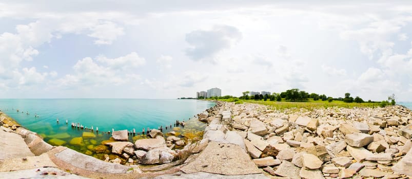 Tranquil lakeside scene with turquoise waters, rugged breakwater, and distant urban skyline - a serene blend of nature and cityscape.