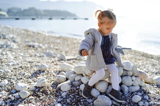 Little girl sits on a pile of stones on a pebble beach by the sea. High quality photo