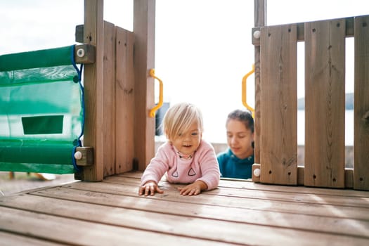 Little girl crawls on her stomach on a wooden deck of a slide against the background of her mother. High quality photo