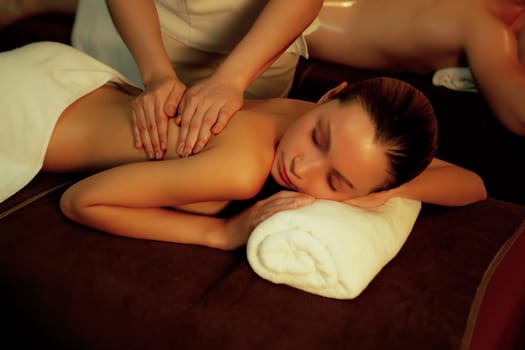 Caucasian couple customer enjoying relaxing anti-stress spa massage and pampering with beauty skin recreation leisure in warm candle lighting ambient salon spa at luxury resort or hotel. Quiescent