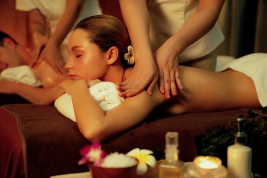 Caucasian couple customer enjoying relaxing anti-stress spa massage and pampering with beauty skin recreation leisure in warm candle lighting ambient salon spa at luxury resort or hotel. Quiescent
