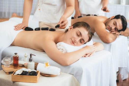 Hot stone massage at spa salon in luxury resort with day light serenity ambient, blissful couple customer enjoying spa basalt stone massage glide over body with soothing warmth. Quiescent