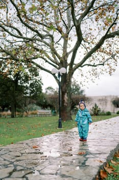 Little girl in overalls stands on a wet paved path in the park. High quality photo