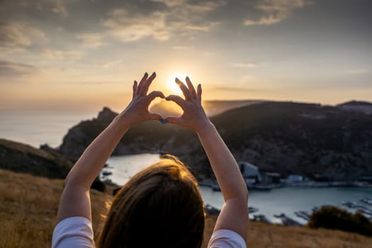 Happy woman on mountain peak, she makes heart shape with hands. Mountain, overlooking sea at sunset. Depicting love shape amidst scenic natural setting