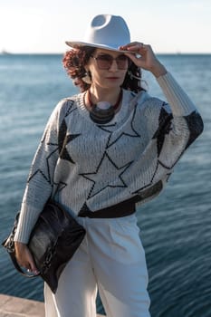 Stylish seashore woman. Fashionable woman in a white hat, white trousers and a light sweater with a black pattern on the background of the sea