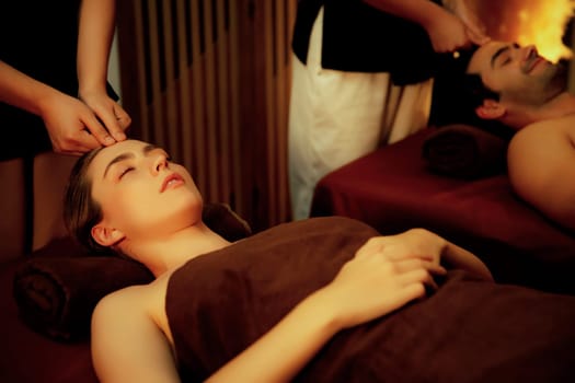 Couple customer enjoying relaxing anti-stress head massage and pampering facial beauty skin recreation leisure in warm candle lighting ambient salon spa in luxury resort or hotel. Quiescent