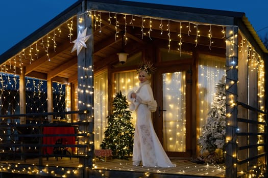 Happy blonde as Snow Maiden in white costume. Wooden house exterior Festive setting. Character with crown creating festive scene