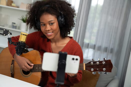 Host channel in musician of young African American playing guitar along with singing, broadcasting on smartphone in studio. Decoration of equipment headsets and recording microphone. Tastemaker.