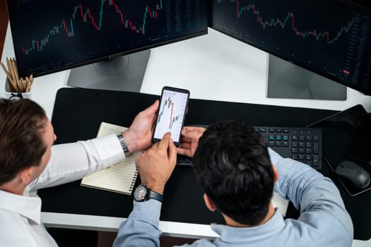 Investor stock officer focusing on dynamic exchange rate database on smartphone, comparing with market dynamic graph with monitor screen. Concept of analysis trading technology investment. Sellable.