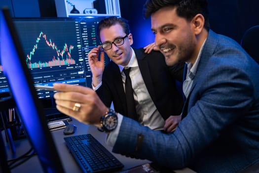 Two stock exchange traders discussing on dynamic successful investment graph, pointing trending of high profit in currency stock in financial data on monitor in neon light at workplace. Sellable.