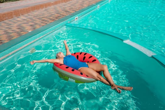 Happy woman in a swimsuit and sunglasses floating on an inflatable ring in the form of a watermelon, in the pool during summer holidays and vacations. Summer concept