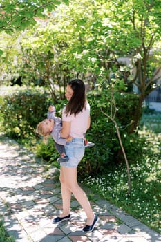 Mom rocks a laughing little girl in her arms in a green garden. Back view. High quality photo