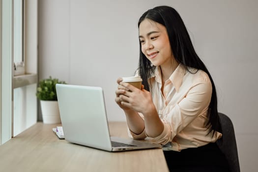 Cheerful Asian female office worker drinking coffee and reading email on her laptop