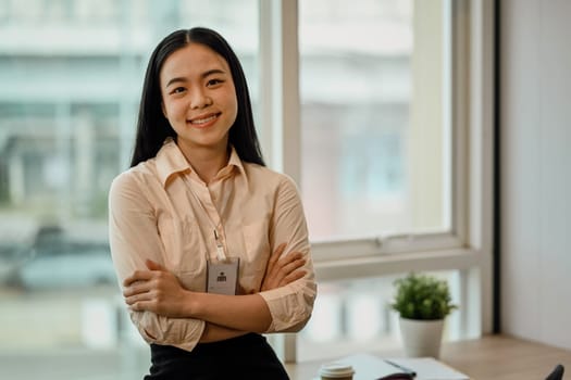 Portrait of a young Asian female employee standing with arms crossed and looks at the camera with a smile