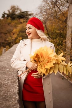 autumn woman in a red beret, a light coat and a red skirt, against the backdrop of an autumn park with yellow leaves in her hands.