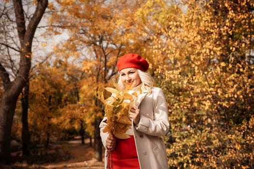 autumn woman in a red beret, a light coat and a red skirt, against the backdrop of an autumn park with yellow leaves in her hands.