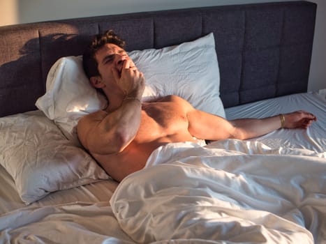 A shirtless man laying on a bed with white sheets, yawning while waking up in the morning
