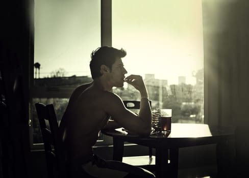 A man sitting at a table in front of a window. Photo of a shirtless young man sitting at a table in front of a window in the morning, eating breakfast and thinking