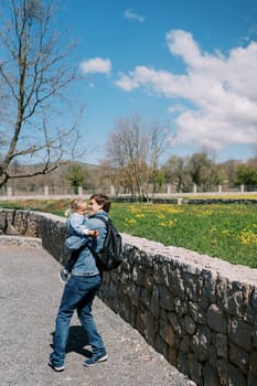 Dad with a little girl in his arms stands near a stone fence on a flowering lawn. High quality photo