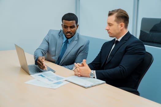 African male leader explain strategy while show solution to professional project manager by pointing at document, using laptop. Skilled business team brainstorming idea. Business meeting. Ornamented.