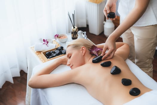 Hot stone massage at spa salon in luxury resort with day light serenity ambient, blissful woman customer enjoying spa basalt stone massage glide over body with soothing warmth. Quiescent