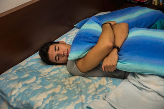 A young man laying on a bed hugging and holding a blue and white pillow. Photo of a young and attractive man lounging on a bed hugging a stylish blue and white pillow